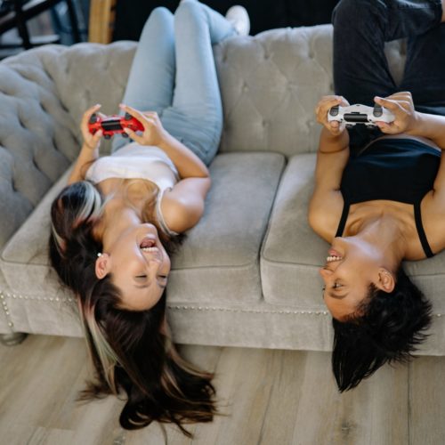 zwei Mädchen liegen auf der Couch und lachen sich an, sie halben einen Controller in der Hand