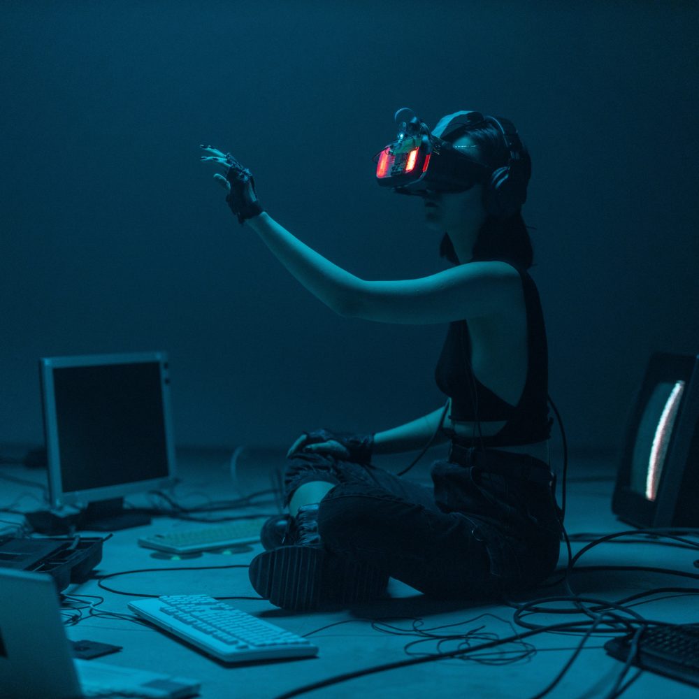 Mädchen mit einer VR-Brille sitzt auf dem Boden umgeben von Bildschirmen. Ihr linker Arm ist gehebt.